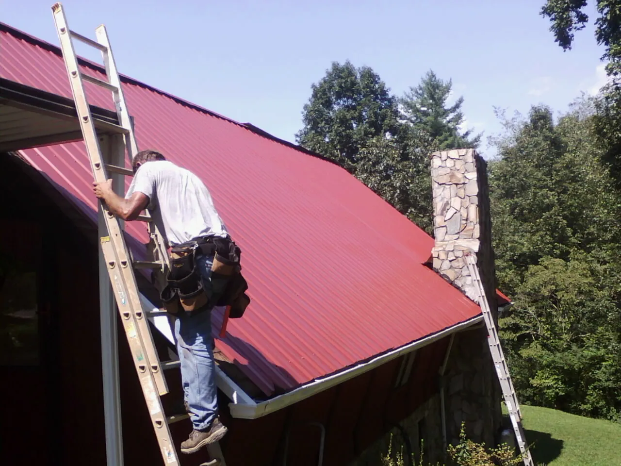 Roofer on a ladder beside a red metal roof and stone chimney — Soffit & Fascia Repair by Roofing Brothers in Terre Haute